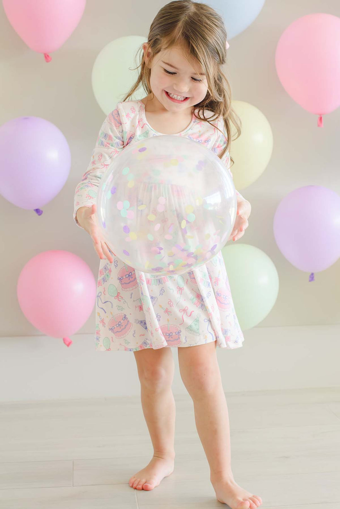 A young girl with light brown hair stands barefoot on a light wood floor, holding a large clear inflatable ball decorated with pastel confetti. She is wearing a Caden Lane long-sleeve, knee-length white dress with a colorful pastel unicorn and cat pattern.