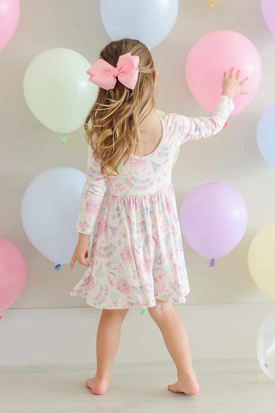 A young girl with long blonde hair and a pink bow is seen from behind, wearing a Caden Lane long-sleeve white dress with a pastel multicolor floral pattern. She is standing barefoot in a room decorated with pastel-colored balloons.