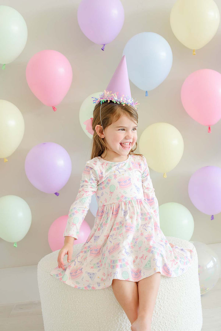 A young girl with a pastel party hat sits on a white textured stool, wearing a Caden Lane long-sleeve pink and blue patterned dress. She is smiling and looking to the side, set against a background of colorful pastel balloons in pink, purple, blue, and yellow.