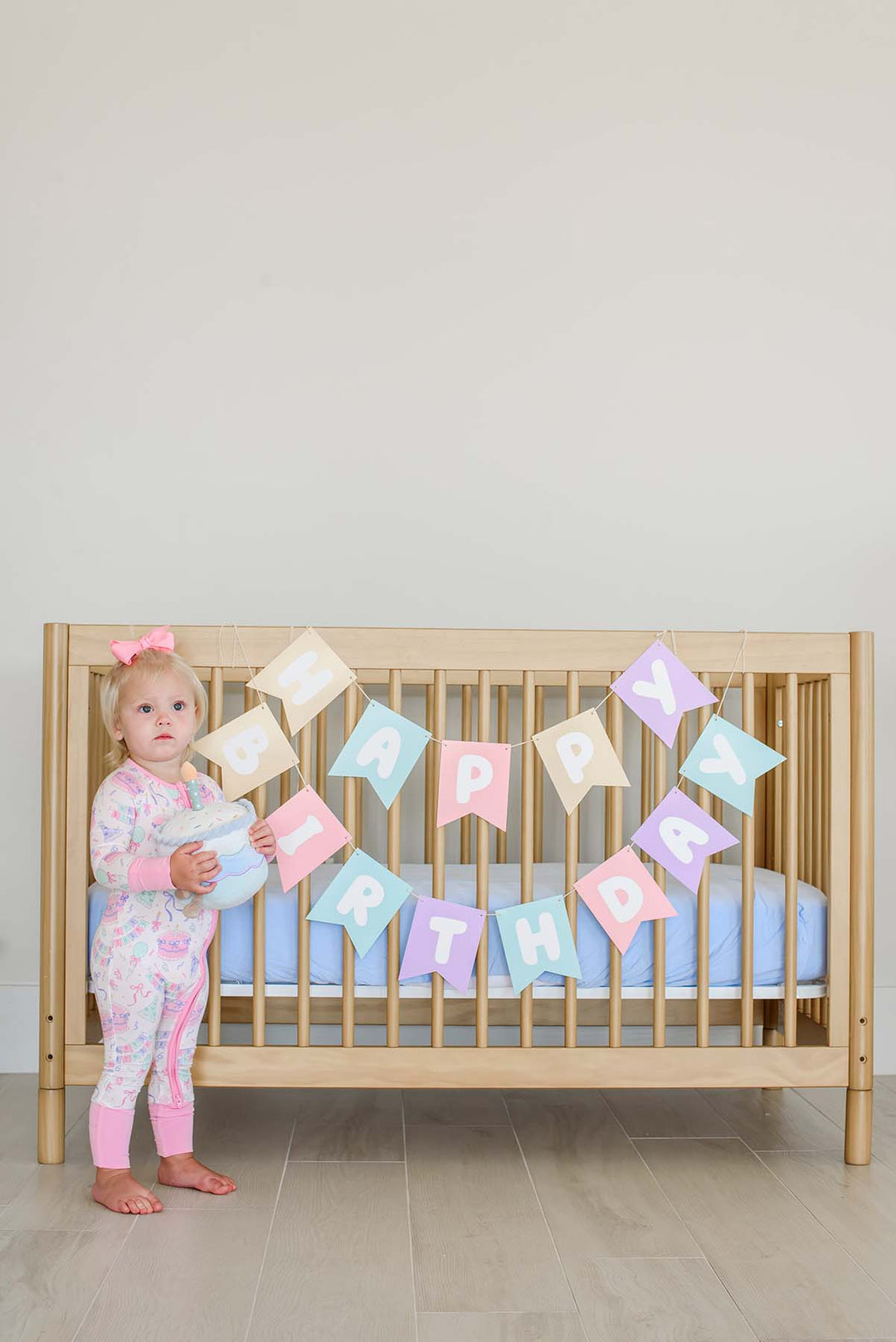 Toddler girl wearing a Caden Lane Birthday Bash zip footie in pink with cake and bow print, holding a plush birthday cake toy beside a wooden crib decorated with a pastel "Happy Birthday" banner.
