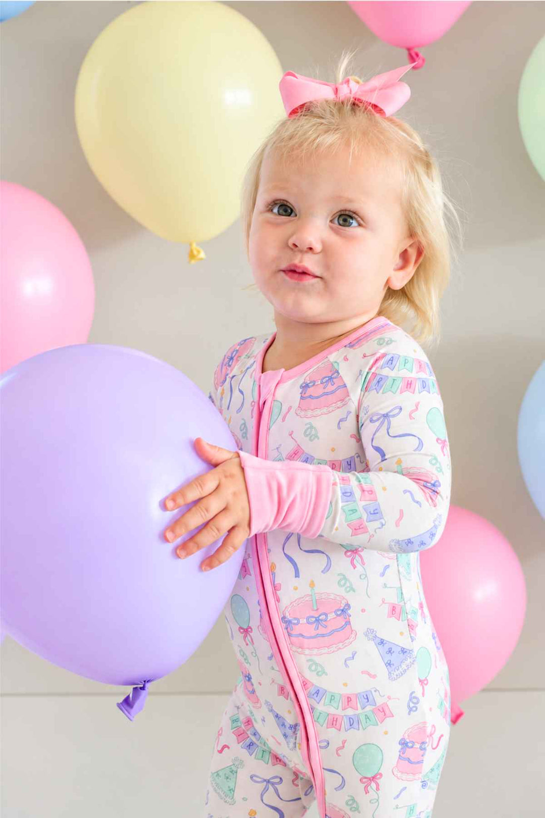 A toddler girl wearing a Caden Lane pink and pastel birthday-themed footed sleeper holds a large lavender balloon, with pink and cream balloons floating in the background.