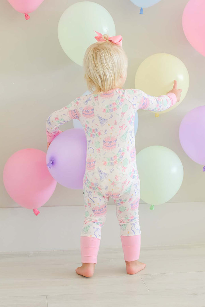 A toddler seen from behind wearing a Caden Lane long-sleeve one-piece pajama with a pastel multicolor cartoon print, standing in front of a wall decorated with pastel balloons.