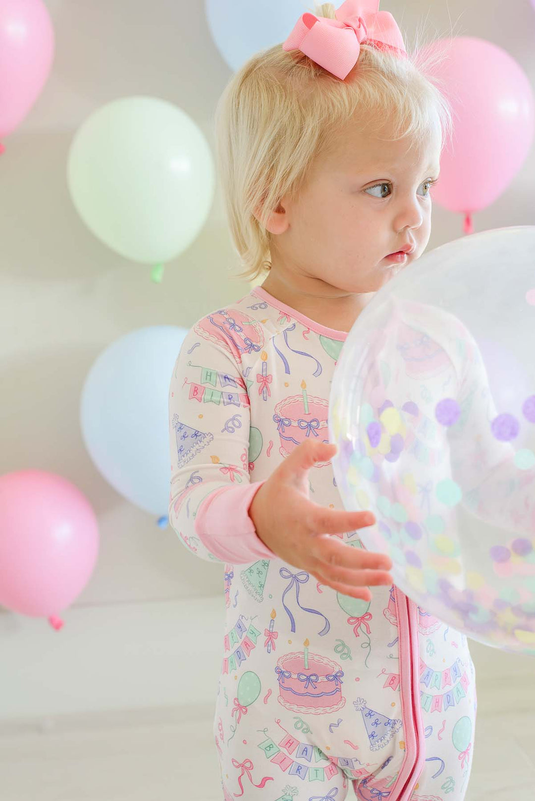 A toddler girl wearing a pink bow in her hair and a light pink long-sleeve footed pajama romper with a Caden Lane colorful birthday print, holding a clear balloon filled with confetti in a room decorated with pastel balloons.