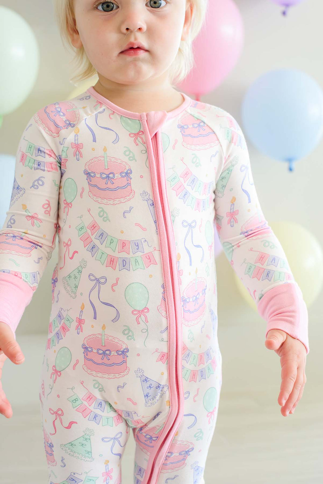 A young child wearing a Caden Lane long-sleeve, one-piece baby jumpsuit with a pink zipper and a colorful birthday print, set against a soft background with pastel balloons.
