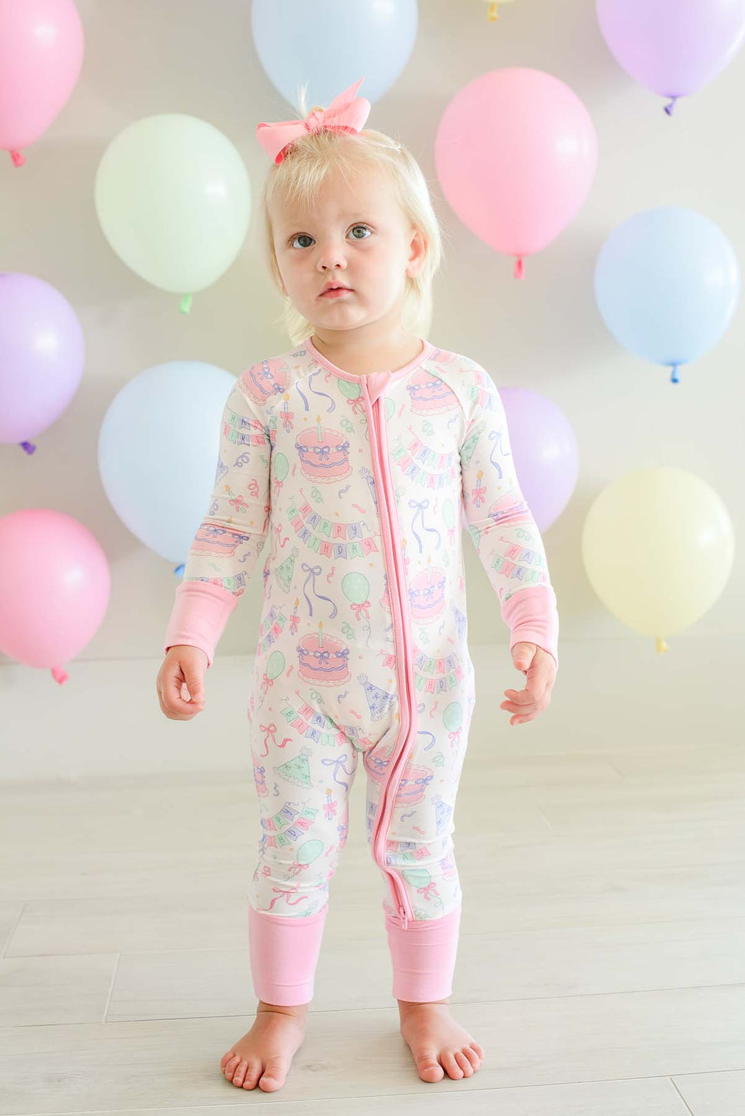 A toddler girl stands barefoot in a room decorated with pastel balloons, wearing a Caden Lane long-sleeve footless pajama romper with a pink zipper and a pastel multicolor print.