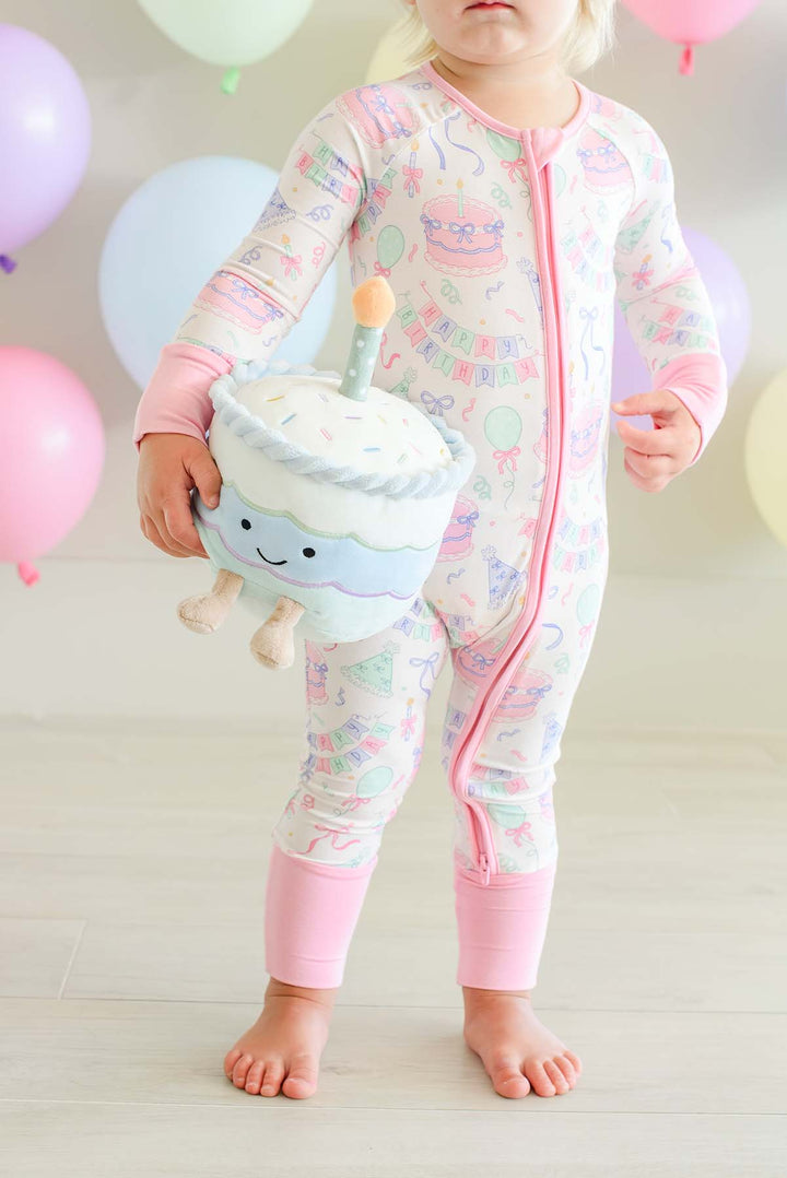A toddler stands barefoot indoors wearing a Caden Lane long-sleeve, one-piece baby pajama with a pink zipper and pastel multicolor birthday-themed print. The pajama features a cupcake character on the chest and matching pink cuffs.