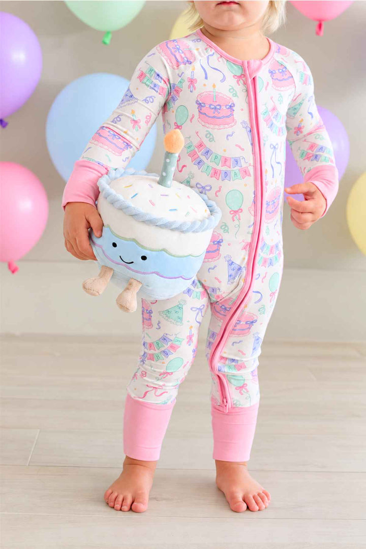 Toddler girl wearing a Caden Lane Birthday Bash zip footie in pink with cake and balloon print, holding a smiling plush birthday cake toy, with pastel balloons in the background.