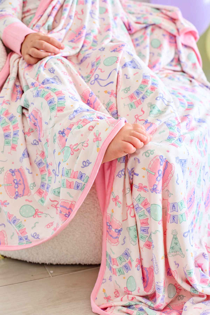 Close-up of the Caden Lane Birthday Bash Pink bamboo blanket featuring birthday cakes, balloons, party hats, confetti and pink trim.