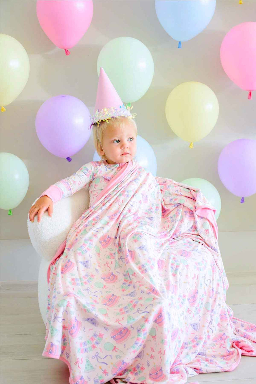 Toddler wearing Caden Lane Birthday Bash Pink bamboo pajamas and wrapped in a matching birthday-print blanket with cakes, balloons and party hats.