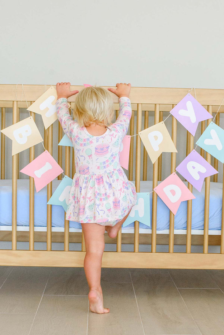 A young toddler stands barefoot in front of a wooden crib, holding up pastel letter flags that spell out 'BIRTHDAY'. The child is wearing a Caden Lane light pink and white long-sleeve dress with a subtle pastel pattern with birthday party motifs.