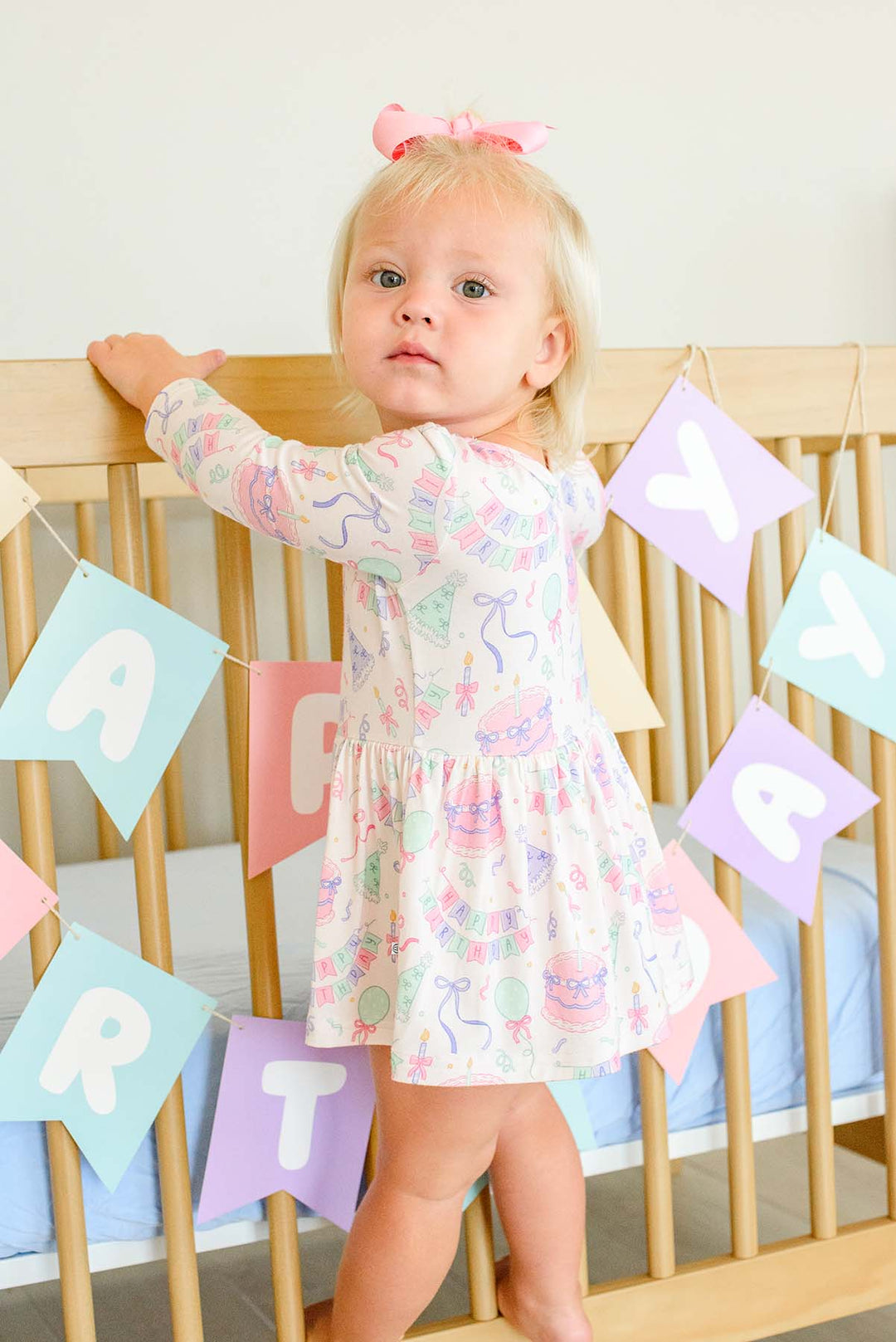 A toddler girl with a pink bow stands in front of a wooden crib wearing a Caden Lane long-sleeve white dress with a colorful pastel party print. Colorful 'PARTY' banner flags hang in the background.