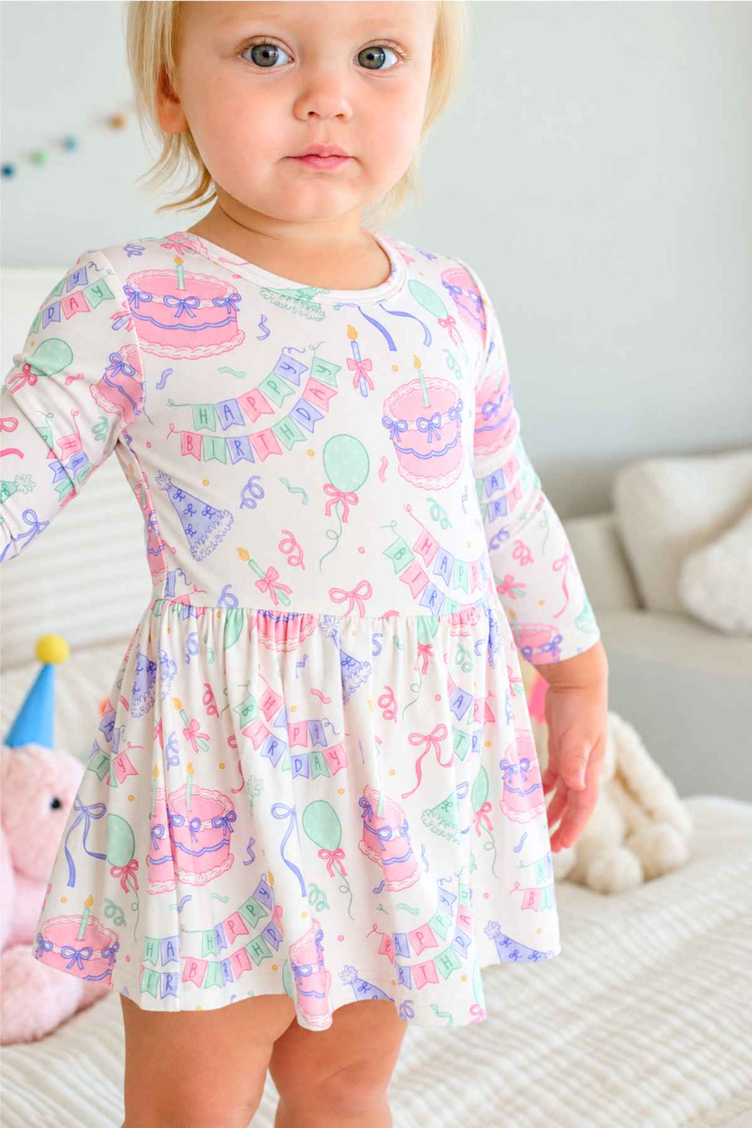 A toddler girl wearing a Caden Lane long-sleeve white dress with a colorful pastel birthday print, standing on a bed in a softly lit room.