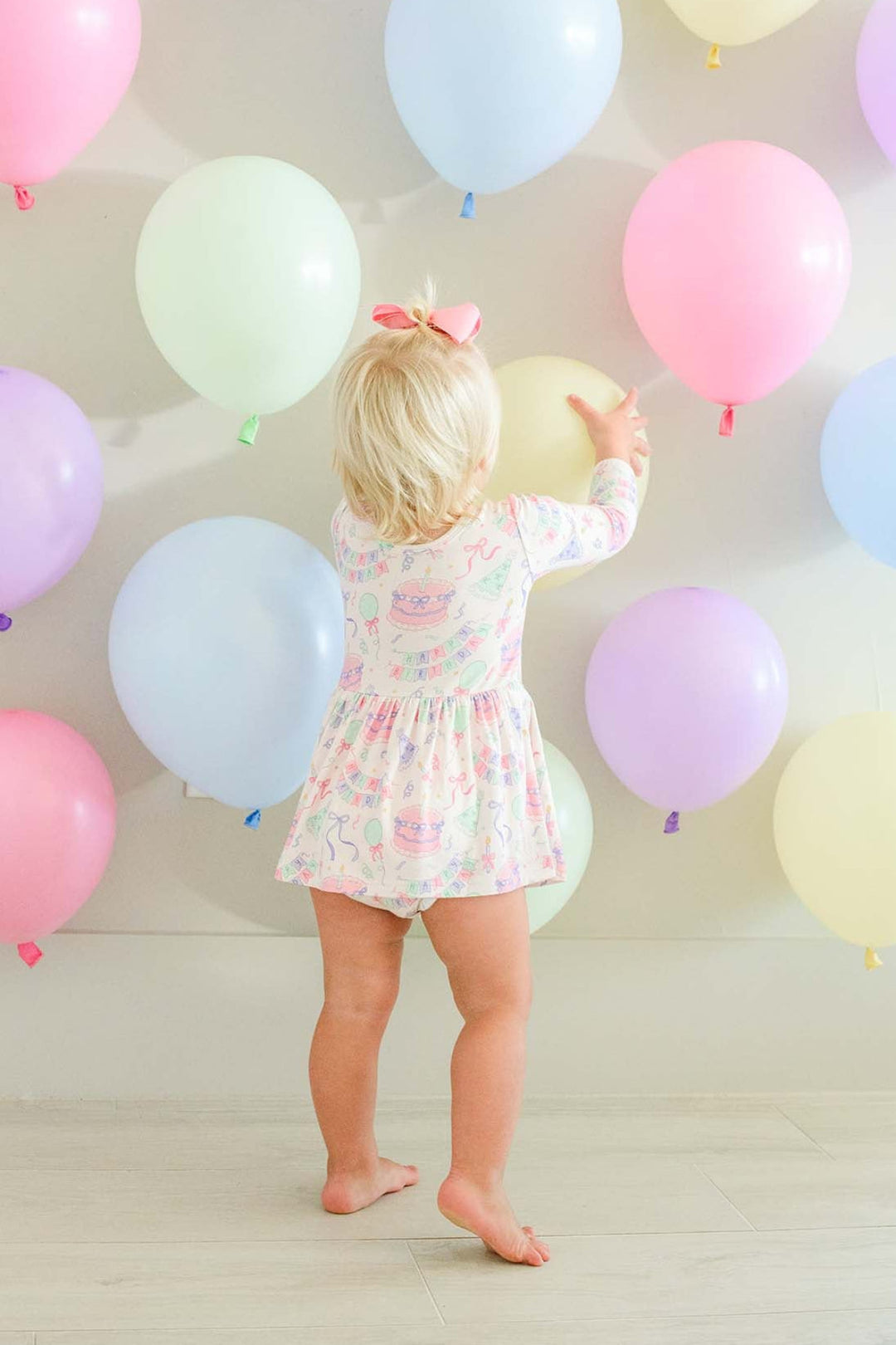 A toddler with blonde hair and a pink bow stands barefoot indoors, facing a wall decorated with pastel balloons in pink, blue, purple, green, and cream. The child is wearing a Caden Lane pink  long-sleeve twirl dress.
