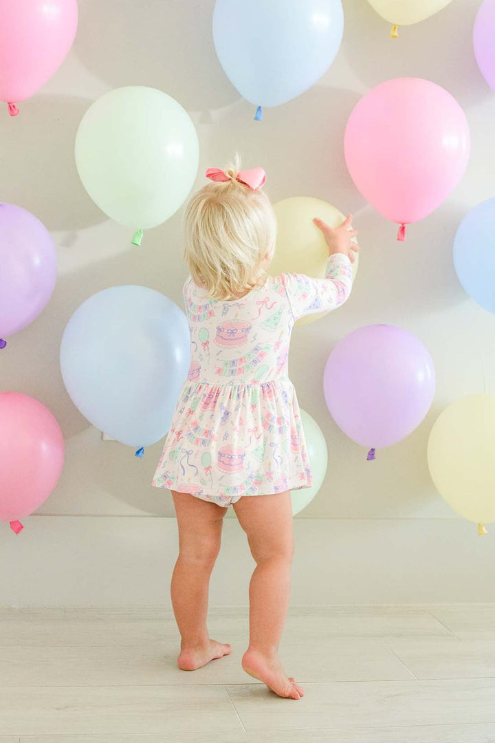 A toddler with blonde hair and a pink bow stands barefoot indoors, facing a wall decorated with pastel balloons in pink, blue, purple, green, and cream. The child is wearing a Caden Lane pink  long-sleeve twirl dress.