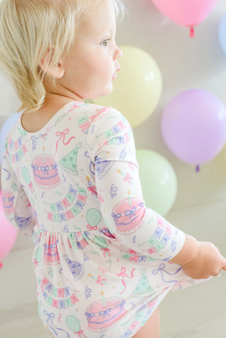 A toddler girl wearing a Caden Lane long-sleeve twirl dress with a colorful birthday print, shown from behind and side profile against a soft background with pastel balloons.