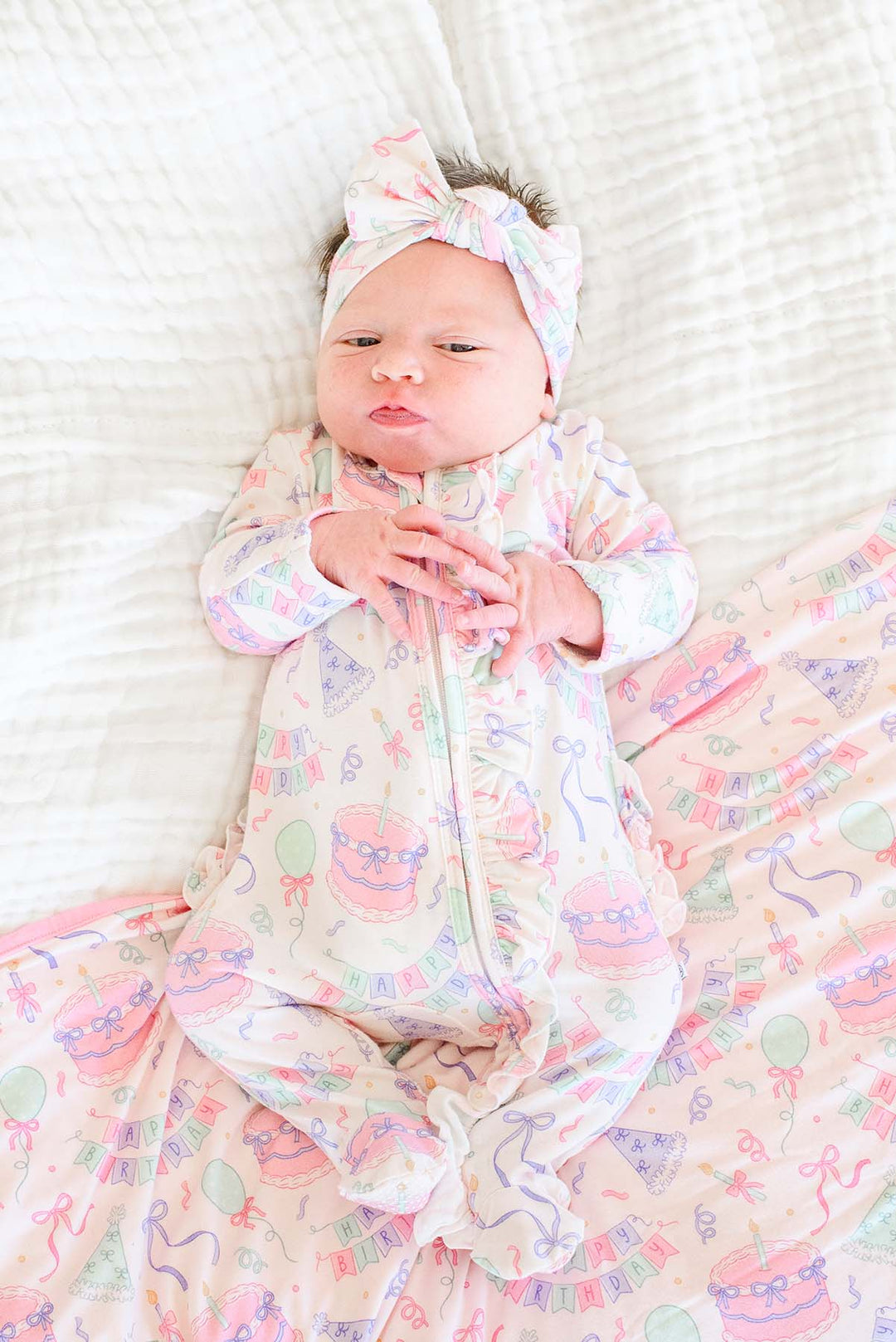 A newborn baby lying on a white textured blanket, wearing a Caden Lane light pink footed sleeper with a matching bow headband and a matching blanket printed with pastel birthday-themed illustrations.