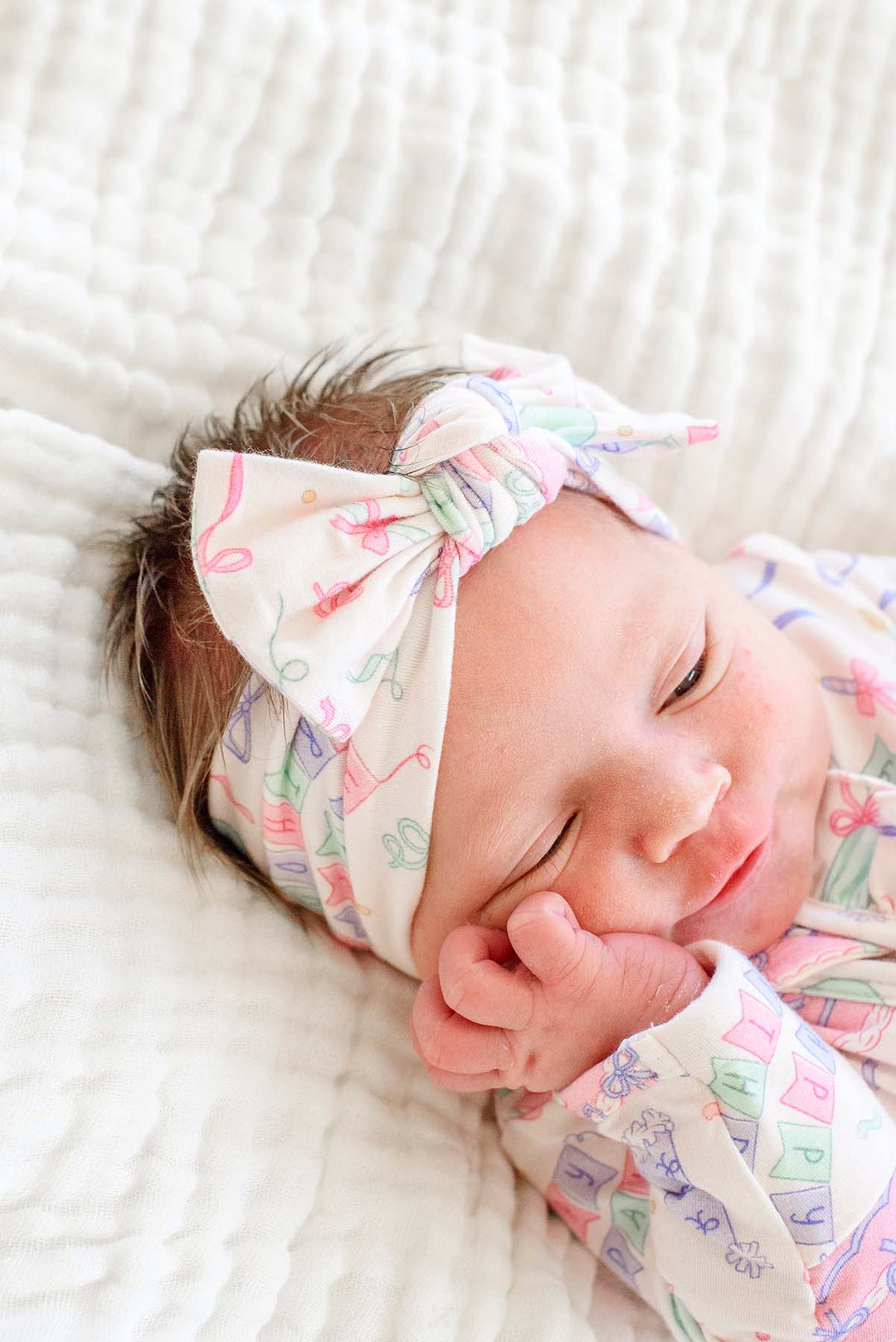 A sleeping newborn baby wearing a Caden Lane birthday print romper and matching bow headband.