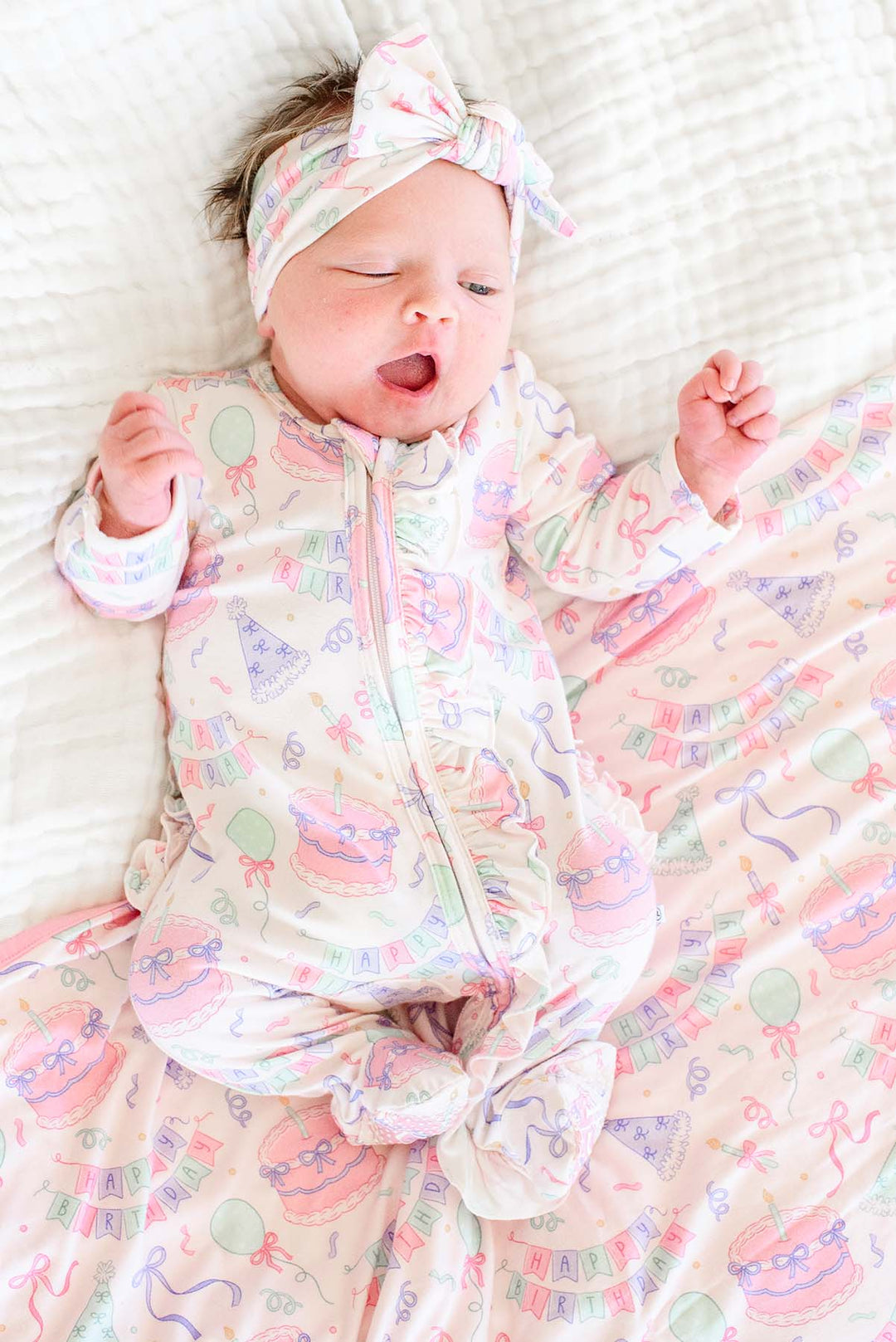 A sleeping newborn baby wearing a Caden Lane pastel ruffle romper and matching bow headband, lying on a matching patterned blanket.