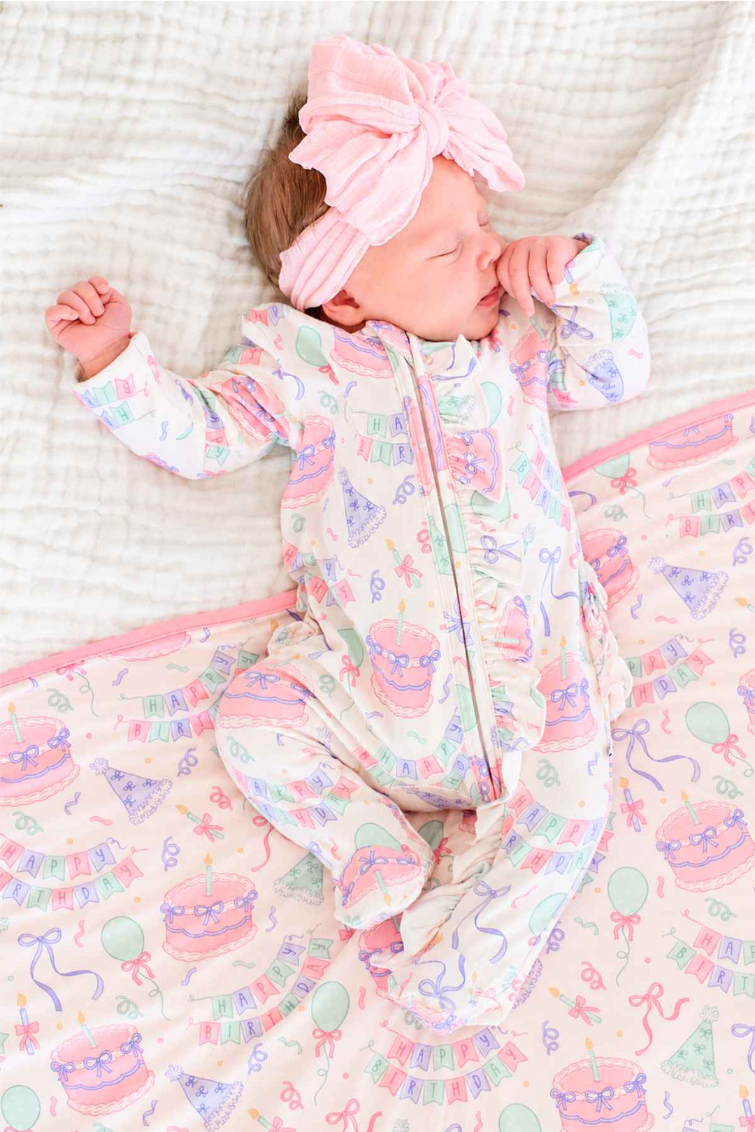 A sleeping newborn baby wearing a light pink bow headband and a Caden Lane white one-piece sleeper with a pastel birthday-themed print, lying on a matching pink and white blanket.