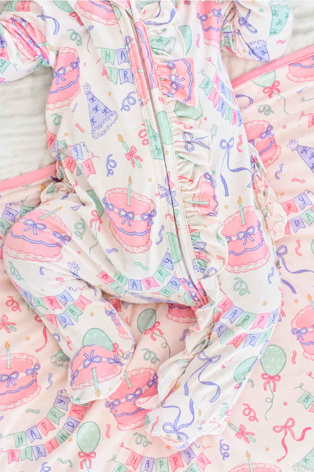 A close-up of a baby's foot and lower leg wearing a Caden Lane light pink footed sleeper with a colorful birthday party print, including cakes, balloons, banners, and bows, lying on a matching pink blanket.