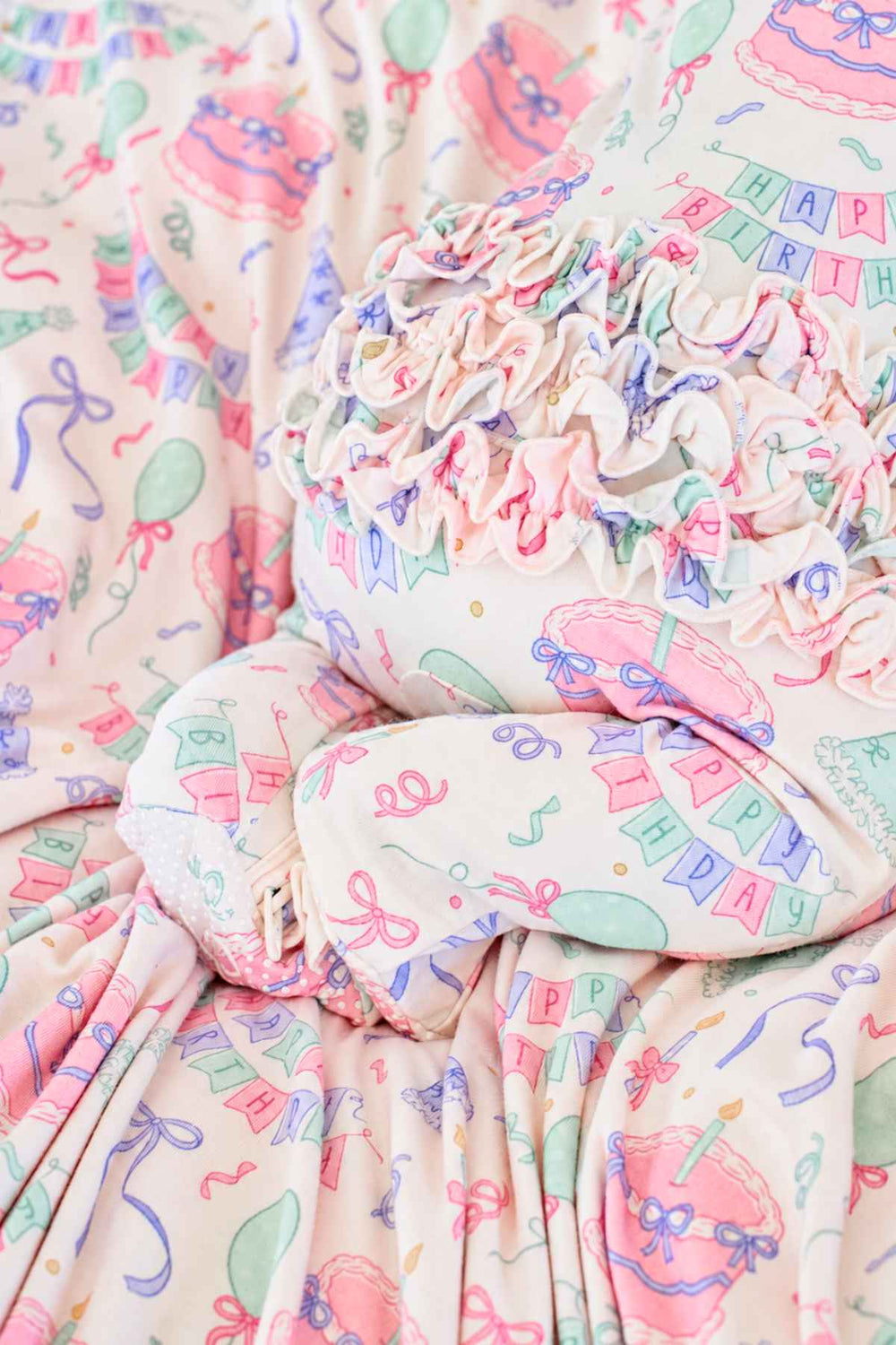A close-up of a baby in a Caden Lane  'Happy Birthday' print ruffle romper, resting on matching patterned blanket.