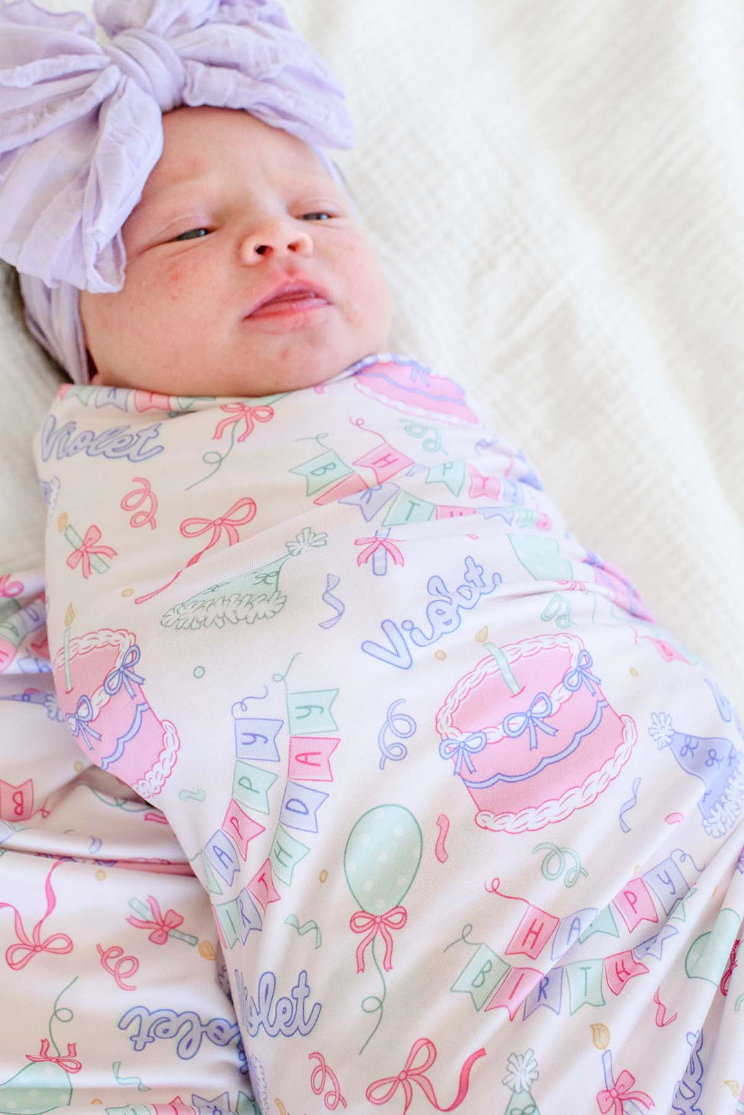 A baby swaddled in a Caden Lane pastel birthday-themed blanket with a matching lavender headband, lying on a white textured blanket.