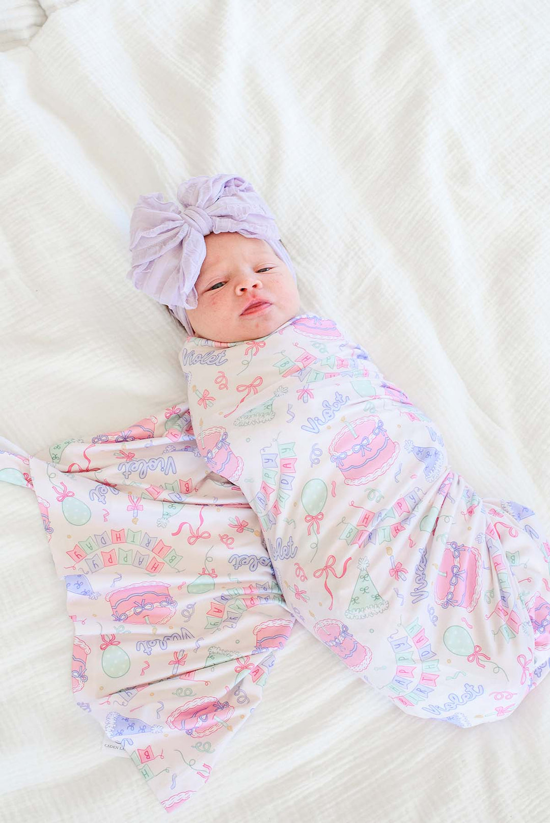 A sleeping newborn baby swaddled in a Caden Lane pastel multicolor blanket with a pink bow headband, lying on a white textured bed.