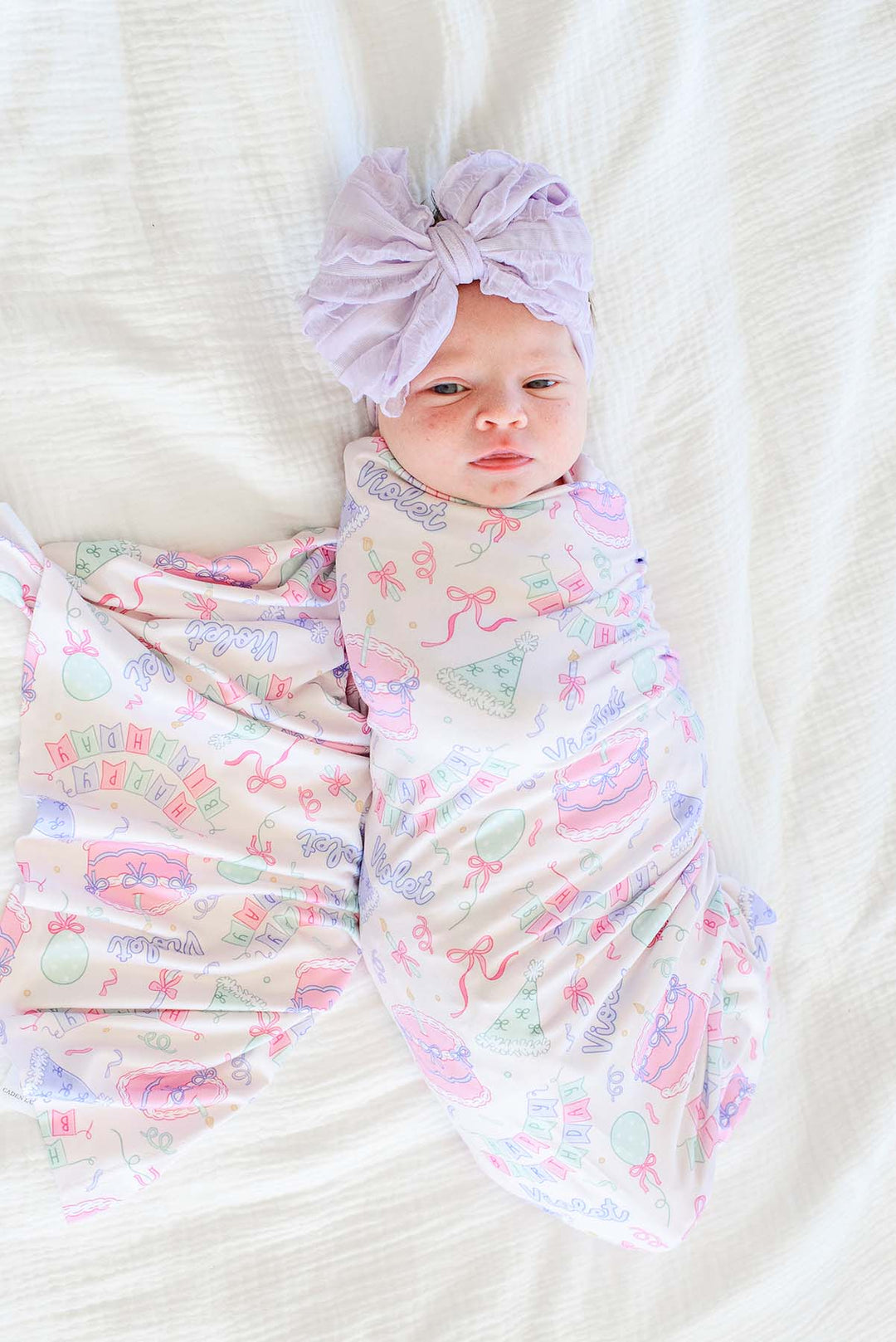 A baby swaddled in a Caden Lane pastel pink and purple patterned blanket with a matching purple bow headband, lying on a white textured blanket.