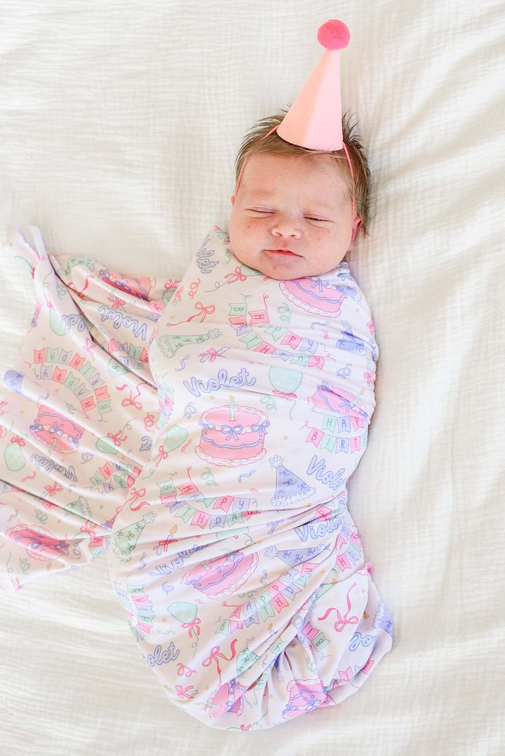 A sleeping newborn baby swaddled in a Caden Lane pastel multicolor blanket with birthday-themed illustrations, wearing a pink party hat, lying on a white textured blanket.