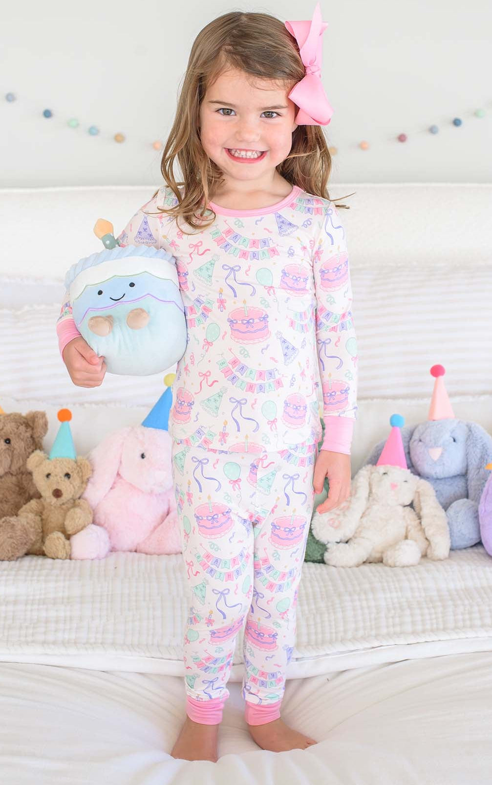 A young girl with a pink bow in her hair stands on a white bed wearing a Caden Lane  long-sleeve pajama set. 