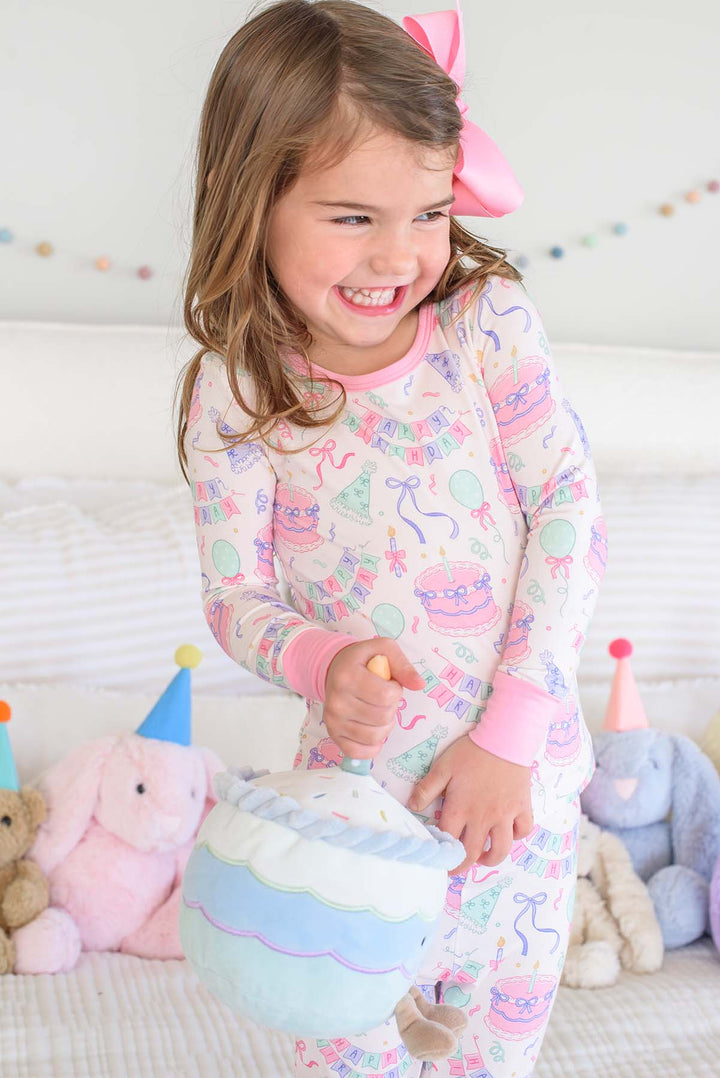 A young girl smiling while wearing a Caden Lane pastel multicolor bunny-themed pajama set with a pink bunny ear headband, standing on a bed with stuffed animals.