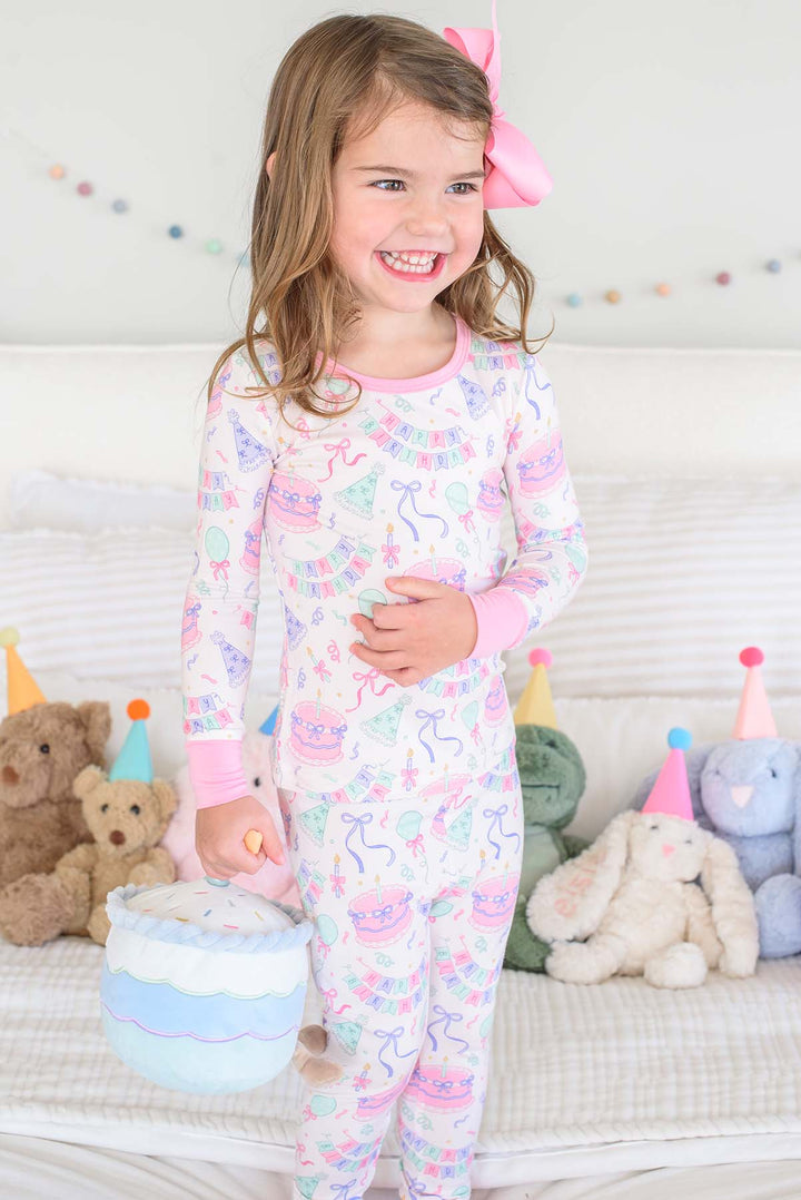 A young girl smiling while wearing a Caden Lane pink and blue patterned long-sleeve pajama set, holding a small blue and white basket in a cozy bedroom setting with stuffed animals on a bed.