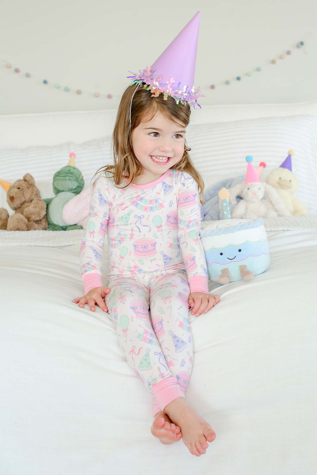 Smiling little girl wearing a Caden Lane Birthday Bash two-piece pajama set in pink with cake and bow print, sitting on a bed in a lavender party hat beside a plush birthday cake toy and stuffed animals in party hats.