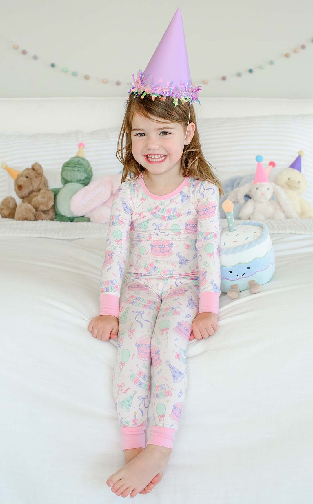 A young girl sitting on a white bed wearing a Caden Lane matching long-sleeve pajama set with a pastel unicorn print and a lavender party hat.