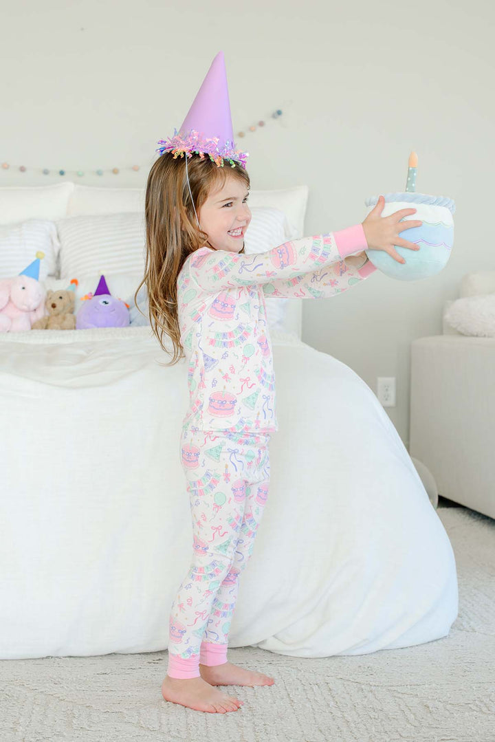 Little girl wearing a Caden Lane Birthday Bash two-piece pajama set in pink with cake and bunting print, wearing a lavender party hat and holding out a plush birthday cake toy with a candle.