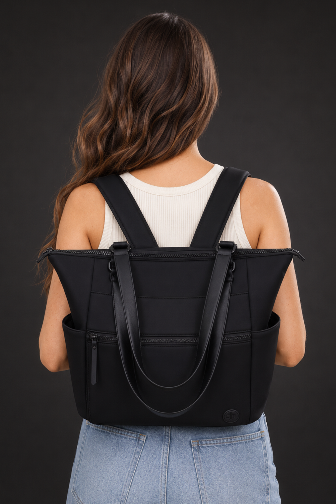 A woman viewed from behind wearing a large black tote backpack with shoulder straps and top handles, paired with a white ribbed tank top and light blue jeans, against a dark gray studio background.
