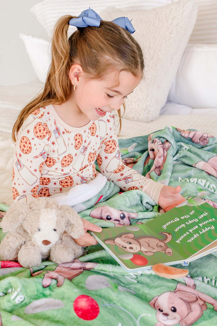 Little girl in cookies-and-milk pajamas sitting on a bed reading the "Little Puppy Plays!" board book, wrapped in a Caden Lane personalized green puppy-print blanket with a plush puppy toy beside her.