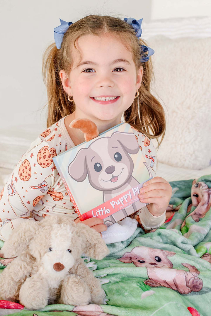 Smiling little girl in pigtails and cookies-and-milk pajamas holding the "Little Puppy Plays!" board book, sitting with a plush puppy toy on a Caden Lane personalized green puppy-print blanket