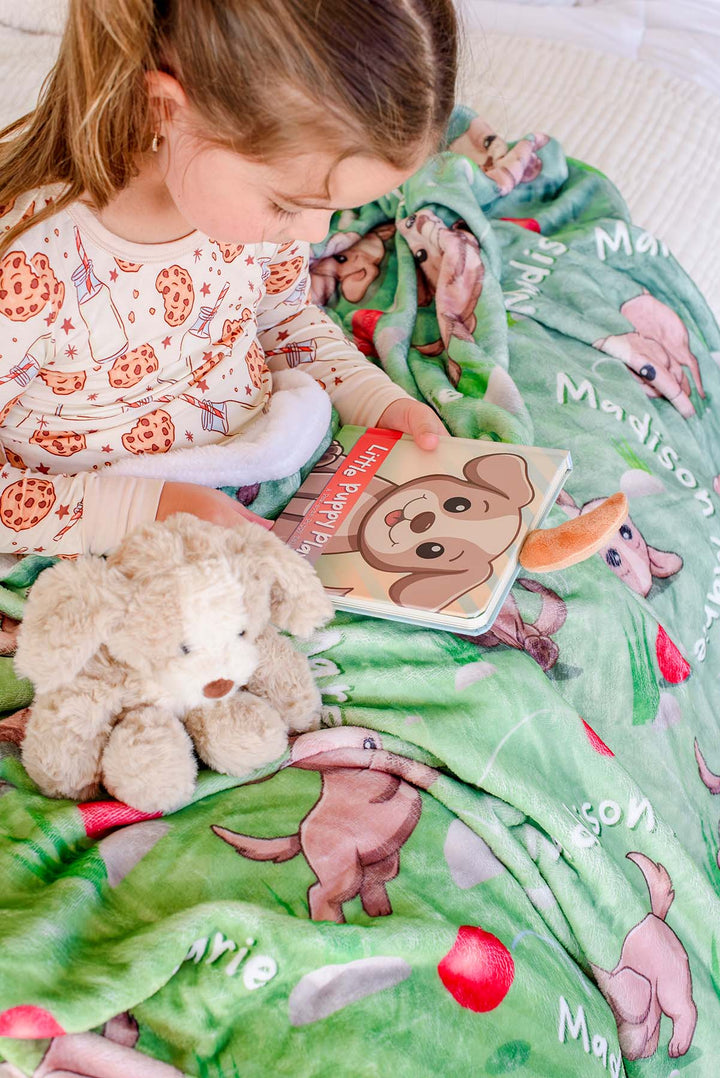 Overhead view of a little girl in cookies-and-milk pajamas reading the "Little Puppy Plays!" board book beside a plush puppy toy, on a Caden Lane personalized green puppy-print blanket with embroidered name visible.