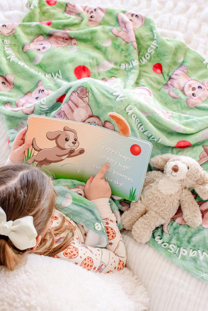 Overhead view of a little girl pointing at a page in the open "Little Puppy Plays!" board book, lying next to a plush puppy toy on a Caden Lane personalized green puppy-print blanket
