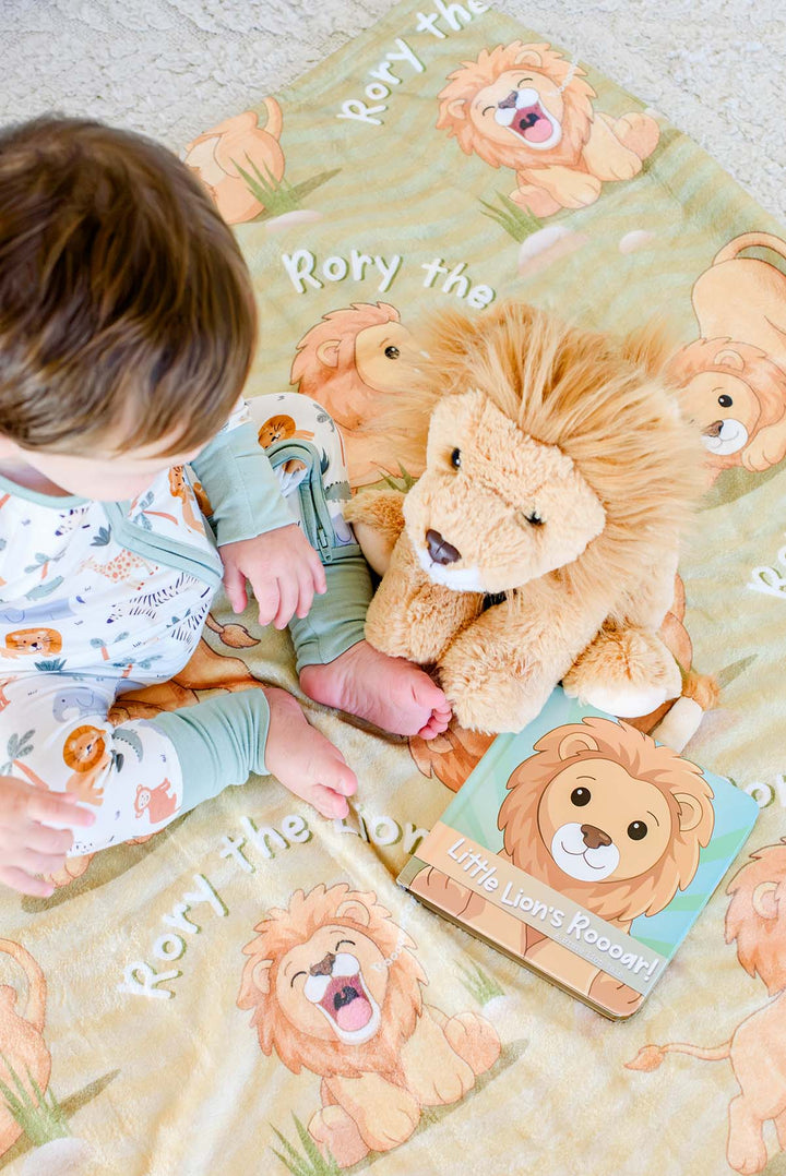 Overhead view of a baby in safari-print pajamas sitting beside a plush lion toy and the "Little Lion's Roooar!" board book on a Caden Lane personalized "Rory the Lion" fleece blanket.