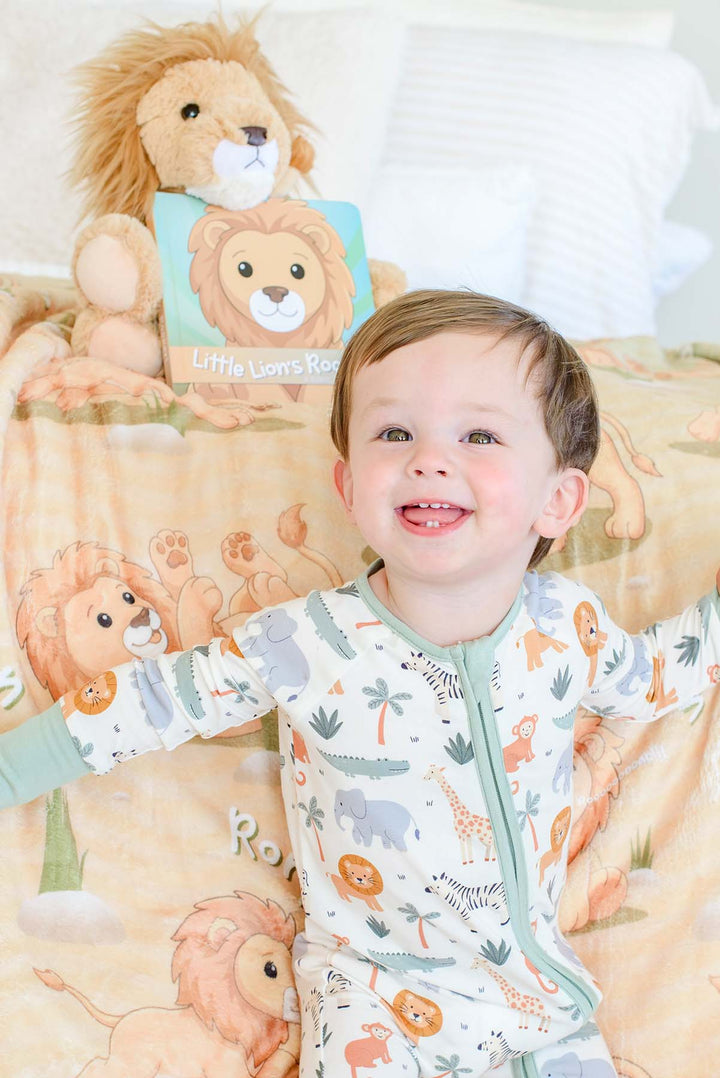 Smiling toddler in safari-print pajamas sitting on a Caden Lane personalized "Rory the Lion" fleece blanket, with a plush lion toy and the "Little Lion's Roooar!" board book beside him