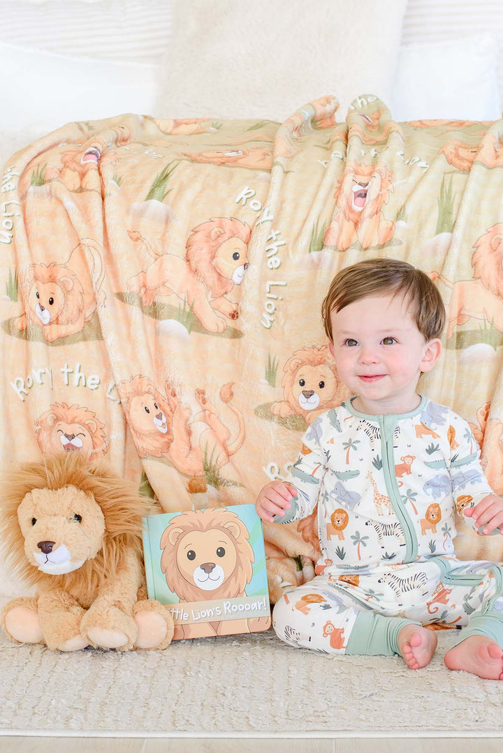Toddler in safari-print pajamas sitting on the floor next to a plush lion toy and the "Little Lion's Roooar!" board book, in front of a draped Caden Lane personalized "Rory the Lion" fleece blanket
