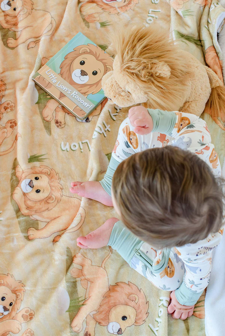Overhead view of a baby in safari-print pajamas crawling on a Caden Lane personalized "Rory the Lion" fleece blanket, next to a plush lion toy and the "Little Lion's Roooar!" board book