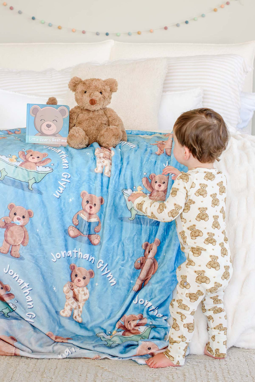 Toddler boy in teddy bear pajamas standing beside a bed with a Caden Lane personalized blue bear-print blanket, a plush teddy bear, and the "Little Bear's Bedtime" board book on top.
