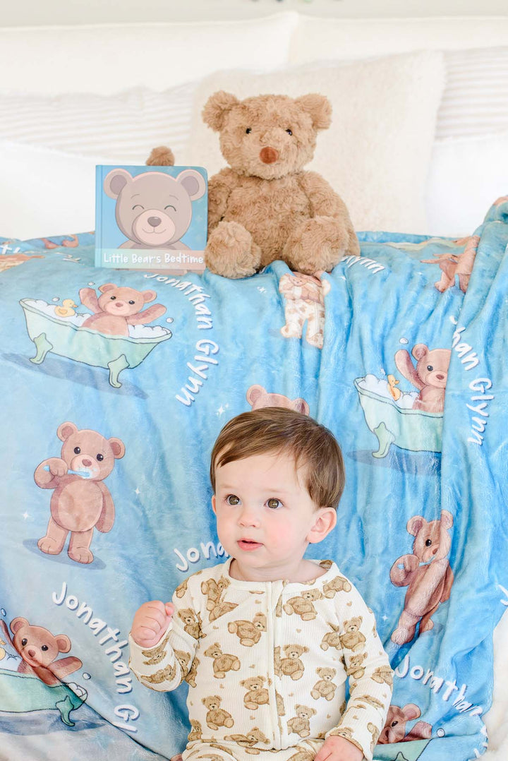 Baby boy in teddy bear pajamas sitting on a bed in front of a Caden Lane personalized blue bear-print blanket, with a plush teddy bear and the "Little Bear's Bedtime" board book behind him.