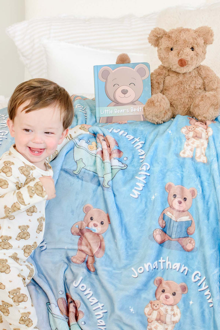 Smiling toddler boy in teddy bear pajamas leaning against a Caden Lane personalized blue bear-print blanket, with a plush teddy bear and the "Little Bear's Bedtime" board book beside him.