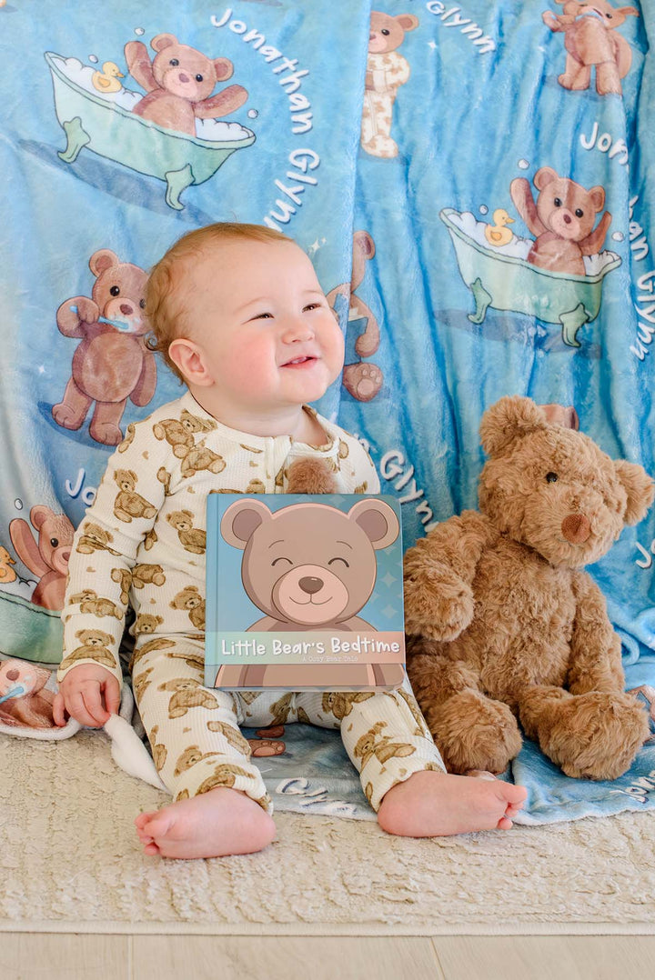 Smiling baby boy in teddy bear pajamas sitting on the floor holding the "Little Bear's Bedtime" board book, next to a plush teddy bear and a Caden Lane personalized blue bear-print blanket.