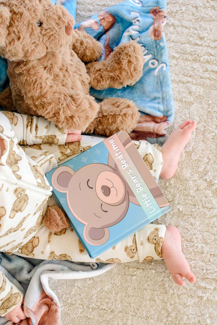 Overhead view of a baby in teddy bear pajamas lying next to a plush teddy bear, the "Little Bear's Bedtime" board book, and a Caden Lane personalized blue bear-print blanket.
