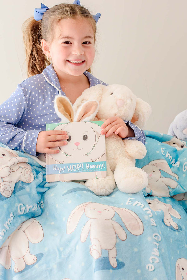 Smiling little girl in blue polka-dot pajamas holding a plush bunny toy and the "Hop! Hop! Bunny!" board book, wrapped in a Caden Lane personalized blue bunny-print blanket.
